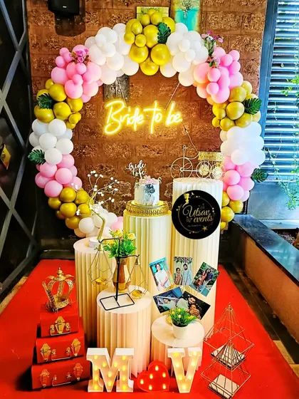 A 'Bride to be' party setup with a colorful balloon ring and a neon sign. The plinths are decorated with photos and other personal touches.