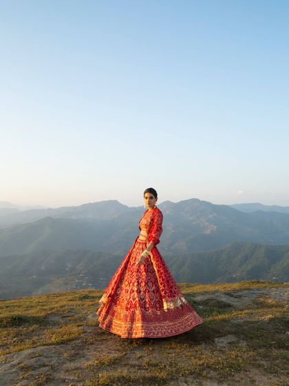 A striking portrait for Frontier Raas, where the model stands on a mountain crest. The styling of the ornate red lehenga against the expansive sky and distant peaks creates a powerful image of a modern, confident bride.