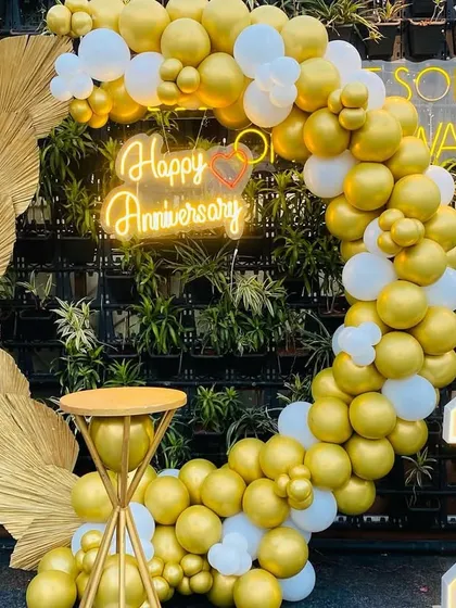 This elegant anniversary decoration features a stunning gold and white balloon ring against a lush green wall. The setup is completed with a "Happy Anniversary" neon sign, large light-up numbers, and dried palm leaf accents for a modern and stylish look.