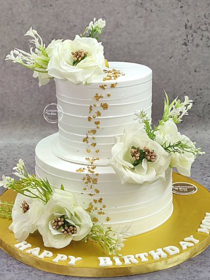 An elegant two-tiered cake for an 80th birthday. The clean white design is adorned with beautiful white flowers and a sprinkle of gold flakes.