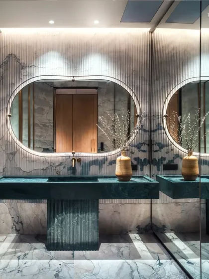 The story of elegance continues in the bathrooms at Palais Royale, where clean lines and precision detailing are paramount. This bathroom features dual vanities in a striking fluted green stone, set against a backdrop of book-matched marble and backlit oval mirrors.