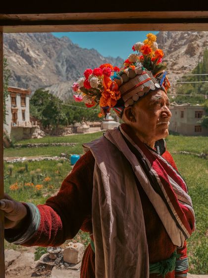 A Brokpa man in traditional red robes and a floral headdress looks out from a doorway. The stunning Himalayan landscape of his village is visible behind him, framing him in his natural environment.
