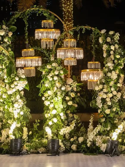 A romantic and elegant photo backdrop for Yashvi and Siddharth's celebration. The setup features arches of white flowers and greenery, accented with modern crystal light fixtures.