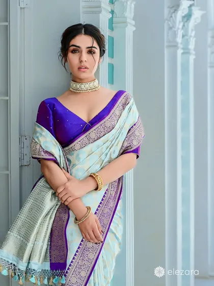 A model for Elezara in a beautiful light blue and purple saree. Her pose against the clean, white pillars creates a look of modern royalty and sophisticated grace.