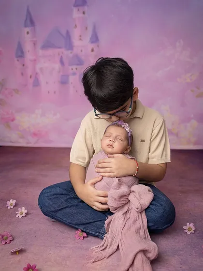 A brother holds his newborn sister in a fairytale setting, with a castle backdrop and soft purple tones.