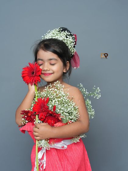 A girl in a flower dress holds a single red gerbera, smelling its fragrance. This tender moment is beautifully captured against a simple grey backdrop.