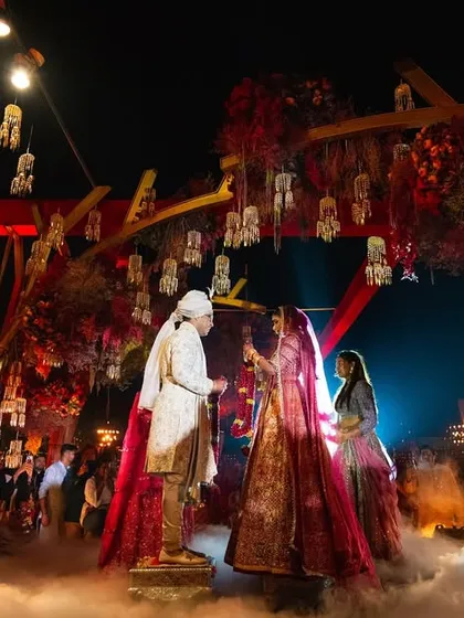 The Varmala exchange under a canopy of red florals and hanging lanterns, surrounded by ethereal smoke. We designed this moment to be the dramatic peak of the ceremony, a fairytale scene where the couple becomes one.