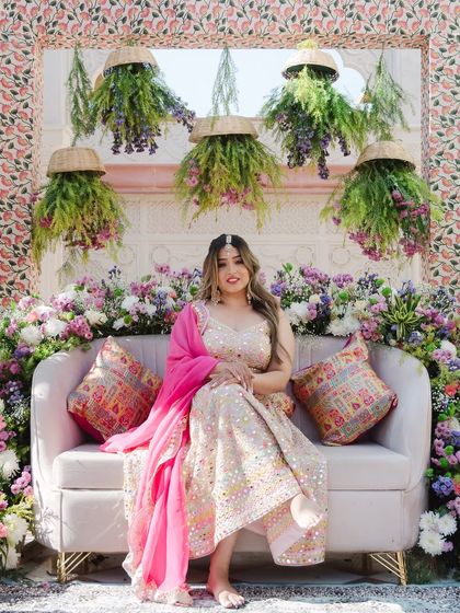 The bride poses on a sofa against a stunning floral backdrop. Her pastel lehenga with a bright pink dupatta creates a vibrant and cheerful Mehendi look.
