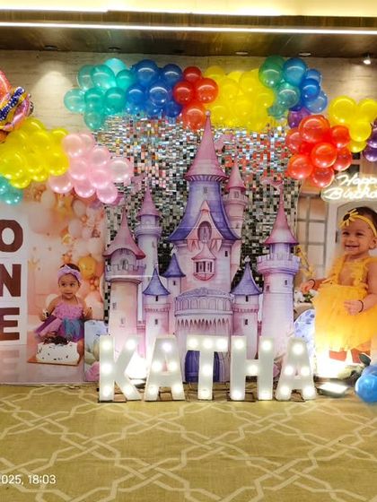 A grand Disney Princess theme for a first birthday. The backdrop features a large castle, cutouts of various princesses like Jasmine and Ariel, and a vibrant, multi-colored balloon arch.
