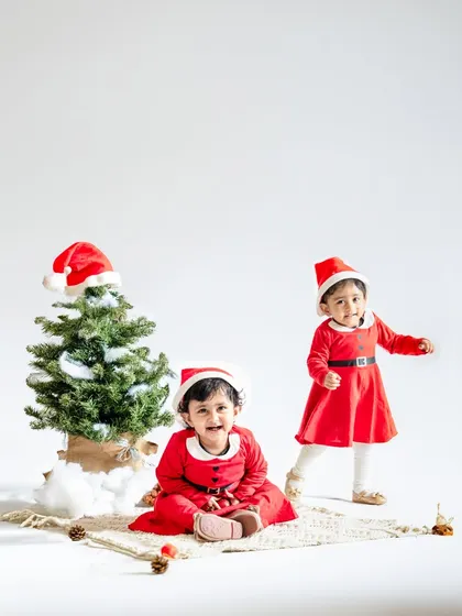 A clean and simple Christmas setup with two sisters in matching Santa dresses. The minimalist white background makes their bright red outfits pop.