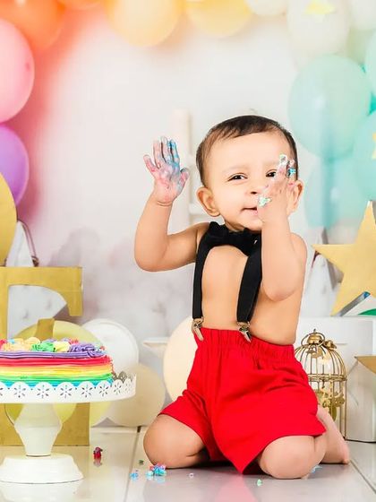 A baby boy with frosting on his hands smiles cheekily at the camera during his rainbow-themed cake smash. A picture of pure, uninhibited joy.