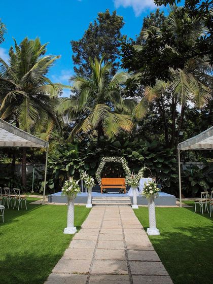 This is the heart of The Farmhouse Collective, set for an intimate wedding. The stone pathway leads to a beautiful stage, surrounded by lush lawns and tropical greenery, creating a perfect natural aisle under a brilliant blue sky.