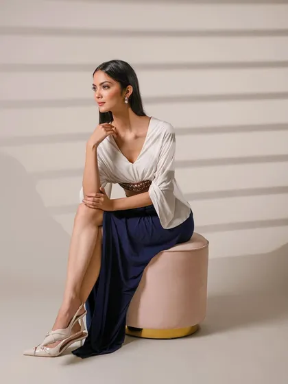 A full-length portrait of a model seated on a pink ottoman, wearing an elegant outfit. The clean studio lighting and shadows create a sophisticated, high-fashion look.