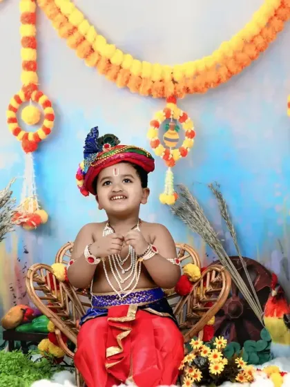 A joyful toddler as Krishna, looking up with a happy expression in a festive setting with marigold decorations and painted pots.