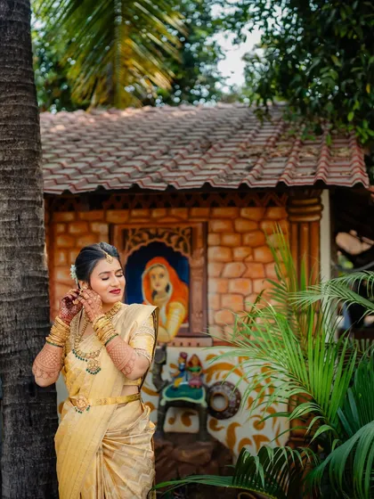 A South Indian bride in a gorgeous golden Kanjivaram saree, posing in a rustic setting. The makeup is soft and luminous.