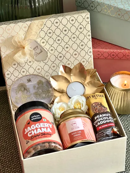 A gift of blessings and good taste. This cream-colored Diwali box includes a Ganesh and Laxmi resin stand, a jar of jaggery chana, chamomile tea, a chocolate ladoo, and a brass lotus tealight holder.