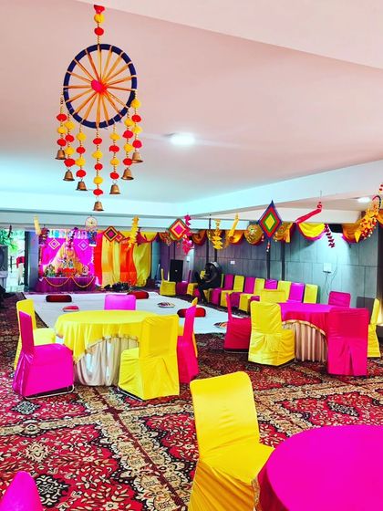 A wide view of a Janmashtami event space, showing the combination of round table seating and floor seating in front of the main stage. The ceiling is decorated with traditional hangings.