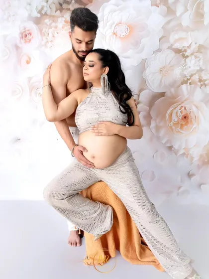 A stylish and intimate couple's portrait. The partner embraces his wife from behind as she sits, both dressed in modern, high-contrast outfits against a floral wall.