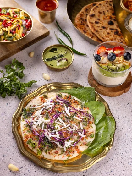 A traditional Indian breakfast spread featuring uttapam, poha, and a fruit parfait. The use of brassware and fresh ingredients gives it an authentic and wholesome feel.
