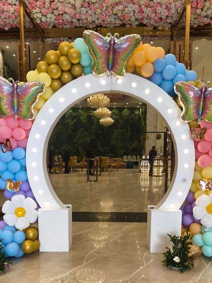 A stunning lighted entrance arch for a grand butterfly-themed party. The arch is decorated with pastel balloons, large foil butterflies, and daisy balloon accents.
