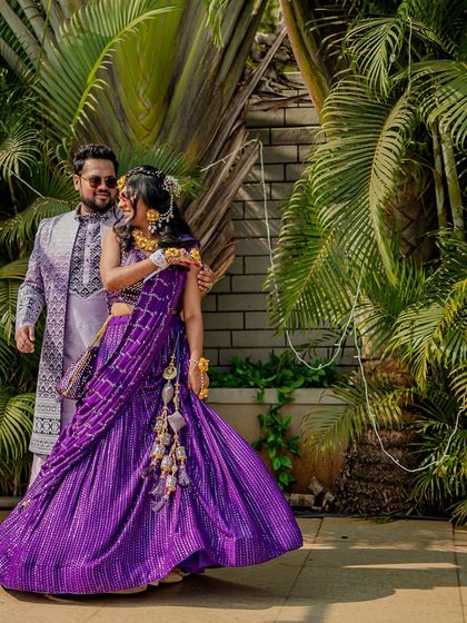 A stunning portrait of the couple in their vibrant Haldi outfits, set against a lush green backdrop. The bride's purple lehenga is a beautiful pop of colour.