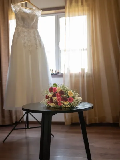 A simple, elegant shot of the wedding dress hanging by the window, with the bridal bouquet waiting on the table. A quiet moment of anticipation.