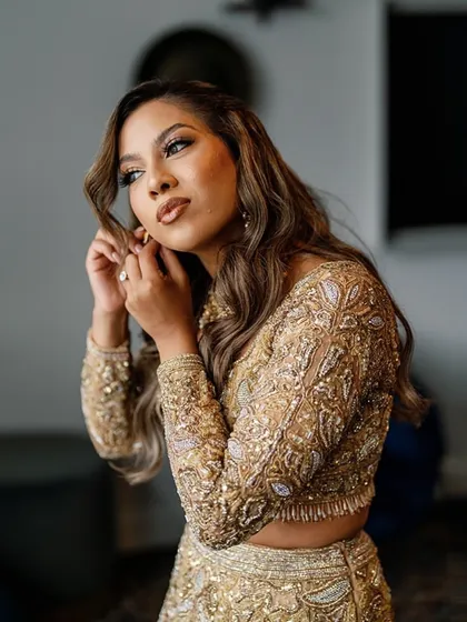 A close-up of the bride getting ready, putting on her earrings. Her soft makeup and wavy hair complement the intricate golden embroidery of her custom lehenga.