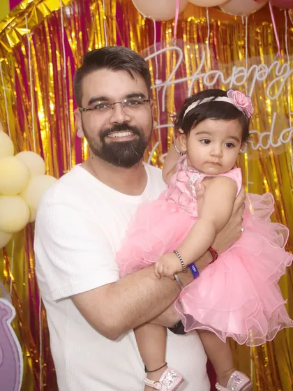 A father and his daughter pose for a photo at her birthday party. The background of pink and gold streamers adds to the celebratory feel of this portrait.