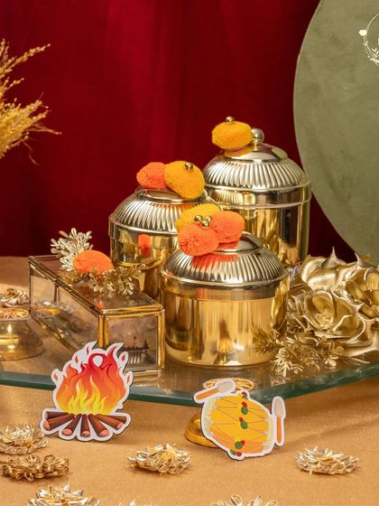 This golden-themed Lohri hamper exudes luxury. It features ornate jars, a glass box for sweets, and festive bonfire cutouts, all arranged on a mirrored tray.