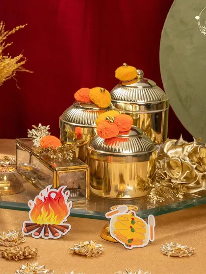 This golden-themed Lohri hamper exudes luxury. It features ornate jars, a glass box for sweets, and festive bonfire cutouts, all arranged on a mirrored tray.