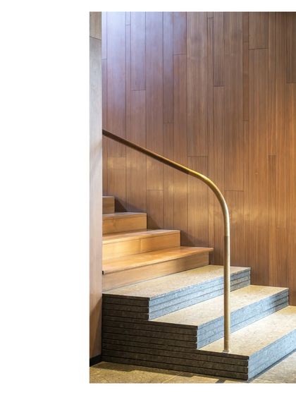 A staircase detail at the Lakeshore house, where stacked stone steps meet a warm wood landing. The aged brass handrail provides a tactile connection point, guiding the transition between materials.