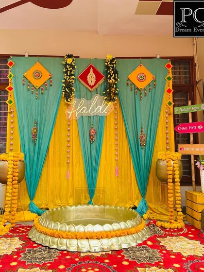 A beautiful and balanced Haldi setup for a home ceremony. I used teal and yellow drapes, traditional hangings, marigold-draped pots, and a central urli to create a festive corner.