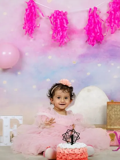 A beautiful portrait from a first birthday session, with the little girl sitting in front of a pastel backdrop with pink balloons and a pretty cake.