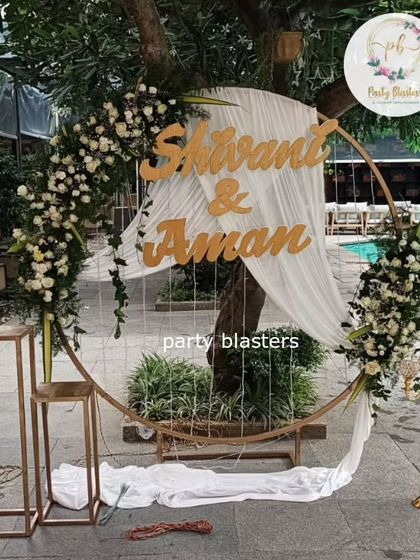 A chic anniversary party setup by the pool. The circular backdrop with the couple's names, draped fabric, and white floral accents creates a modern and romantic photo opportunity.