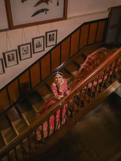 A high-angle shot that showcases the magnificent trail of the bride's lehenga as she poses on the grand wooden staircase. This perspective adds a touch of drama and elegance.