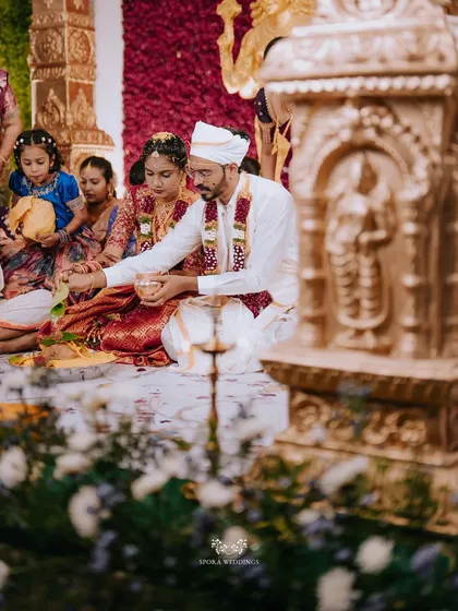 The couple performing a ritual during their wedding ceremony, set against a backdrop of red roses and golden idols.