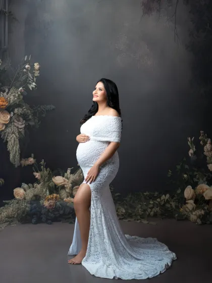 A portrait of grace. Set against a moody, floral backdrop, the expectant mother in a white lace gown looks upward, creating a hopeful and elegant image.