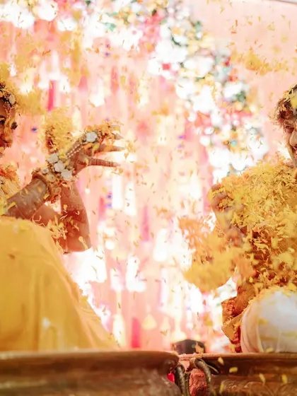 A beautiful, playful moment from a Haldi ceremony. The couple is covered in turmeric and flower petals, sharing a laugh in front of a delicate backdrop of pastel florals and hanging crystals.