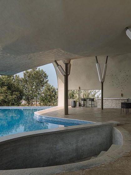 From inside the semi-open lounge, the curved roof elegantly frames the pool and the trees. The support columns are designed to resemble branching trees, reinforcing the connection between the architectural elements and the natural world.
