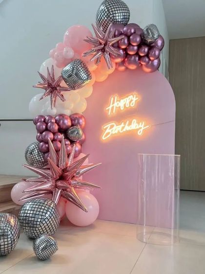 A close-up of a trendy disco-themed birthday decoration. This setup features a pink arched backdrop, a neon sign, and a garland of pink and chrome balloons mixed with starburst and disco ball foils.