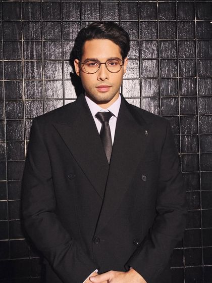 A close-up portrait of Siddhant Chaturvedi. The addition of clear-framed glasses adds a scholarly, intellectual touch to the formal black suit, showing how accessories can define a look.