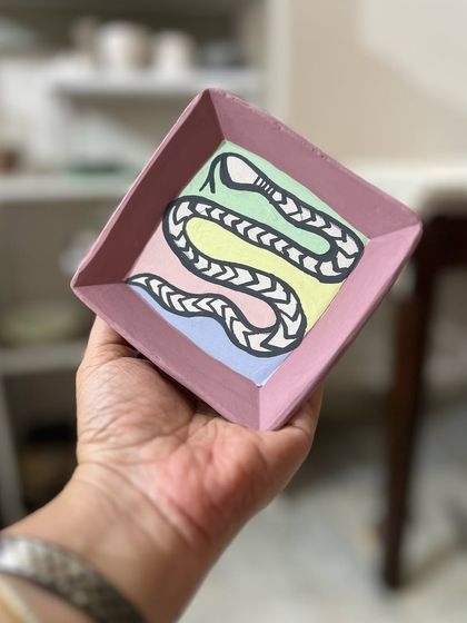 A folksy, hand-painted snake plate with a pastel, color-blocked background. This piece combines traditional motifs with a modern color palette, creating a unique trinket dish.
