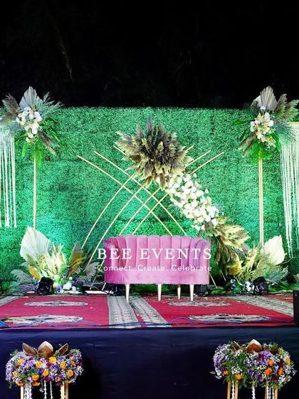 This reception stage combines rustic charm with modern elegance. A lush green wall serves as the backdrop, accented with boho-style pampas grass, macrame hangings, and a vibrant pink sofa for a unique and stylish look.