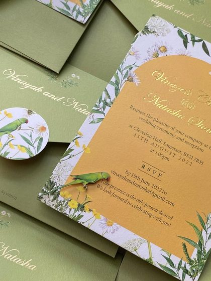 The complete invitation suite for the UK wedding, showing the main card, the custom envelope, and a sticker featuring the parrot motif. The design is a celebration of nature and shared heritage.