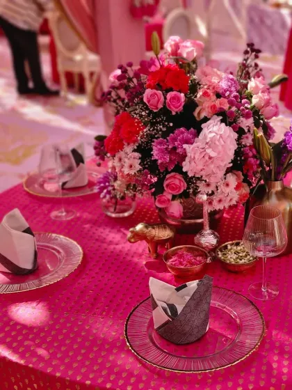 A tablescape from a vibrant Anand Karaj lunch. The bright pink tablecloth with gold motifs is paired with elegant glass plates and a stunning centerpiece of pink and purple flowers.
