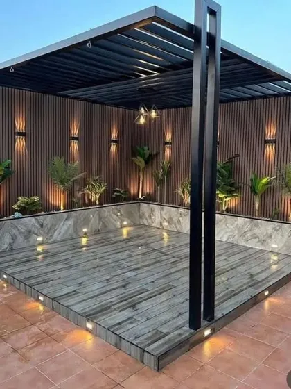 A stylish pergola with a black metal frame, wood-paneled back wall, and a marble-look tiled floor. The space is illuminated with both in-floor uplights and wall sconces for a dramatic effect.