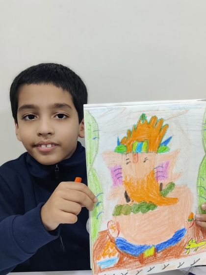 A young boy proudly holds up his colourful drawing of Lord Ganesha, created with oil pastels. His interpretation features a bright orange Ganesha with a decorative crown and a friendly mouse companion.