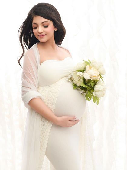 A close-up shot highlighting the baby bump, with the mother-to-be holding a bouquet of white peonies. The bright, white-on-white styling creates a pure and delicate look.