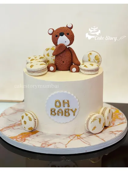 A chic and modern 'Oh Baby' shower cake. The simple white cake is decorated with a fondant teddy bear and elegant macarons with gold leaf details.