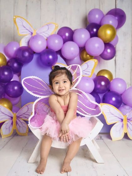 A sweet toddler dressed as a butterfly, smiling brightly for her milestone photoshoot. The purple and gold balloon arch and butterfly cutouts create a magical, whimsical setting.